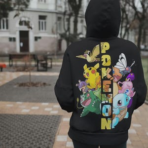May include: Black hooded sweatshirt featuring a colorful graphic of various cartoon characters and the word "POKEMON" in yellow. The design includes a variety of creatures in vibrant colors. The sweatshirt is worn outdoors.