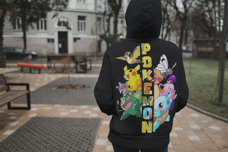 May include: Black hooded sweatshirt featuring a colorful graphic of various cartoon characters and the word "POKEMON" in yellow. The design includes a variety of creatures in vibrant colors. The sweatshirt is worn outdoors.