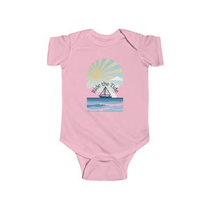 May include: A light pink baby bodysuit with a graphic of a sailboat on the ocean under a sun and the text "Ride the Tide". The design features blue, yellow, and white colours.