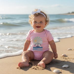 May include: A baby in a pink bodysuit with a nautical design sits on a sandy beach. The bodysuit features a sailboat, sun, and the text "Ride the Tide". The baby wears a white bow headband and smiles. The sea is in the background.