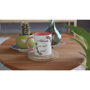 Op de afbeelding: Een witte keramische mok met een roze binnenkant en oor, met de tekst "Atlanta is not real" omringd door een bloemenkrans. De mok staat op een houten onderzetter, met een cactus en een decoratieve vaas op de achtergrond.