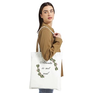 Op de afbeelding: Witte draagtas met de tekst "Atlanta is not real" in een sierlijk lettertype, omgeven door een bloemenkrans. De tas wordt gedragen door een persoon die een bruine jas draagt.