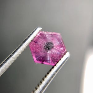 Op de afbeelding: Een gefacetteerde, zeshoekige robijn edelsteen, vastgehouden door een zilveren pincet. De edelsteen is helderroze met een donker centrum en zichtbare interne structuren. De pincet is scherp in focus tegen een vage grijze achtergrond.