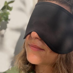 K&ouml;nnte beinhalten: Eine schwarze Schlafmaske bedeckt die Augen und die Nase. Die Maske besteht aus einem weichen, undurchsichtigen Material. Die Person, die die Maske tr&auml;gt, l&auml;chelt. Der Hintergrund ist eine wei&szlig;e Wand mit Schatten und einer gr&uuml;nen Pflanze.