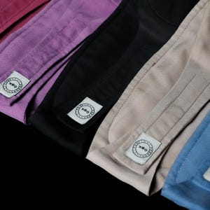 K&ouml;nnte beinhalten: Ein Set aus f&uuml;nf Stoffstirnb&auml;ndern in den Farben Burgunderrot, Lila, Schwarz, Beige und Blau. Jedes Stirnband hat ein rechteckiges wei&szlig;es Stoffetikett mit einem kreisf&ouml;rmigen Logo, einer kleinen Grafik und dem Text "THE SPA HEADBAND" in Schwarz.