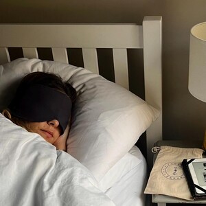 Pu&ograve; includere: Una mascherina per dormire grigio scuro indossata sugli occhi, appoggiata su un cuscino bianco. Accanto al letto, una pochette beige con coulisse presenta un logo circolare con il testo "COSE LONDON" e un piccolo simbolo grafico, accanto a un dispositivo elettronico nero.