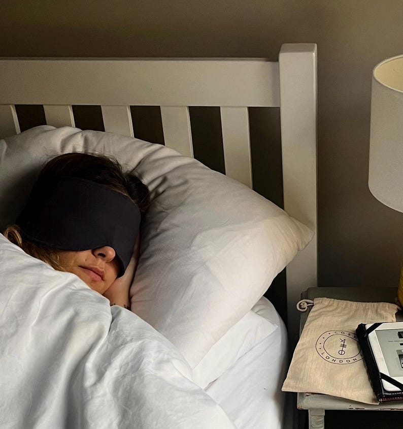 Pu&ograve; includere: Una mascherina per dormire grigio scuro indossata sugli occhi, appoggiata su un cuscino bianco. Accanto al letto, una pochette beige con coulisse presenta un logo circolare con il testo "COSE LONDON" e un piccolo simbolo grafico, accanto a un dispositivo elettronico nero.