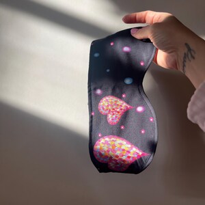 May include: A dark blue sleep mask featuring two heart shapes with colorful, speckled patterns. The mask has a dark background with small, light-colored dots. The mask is held up by a hand.