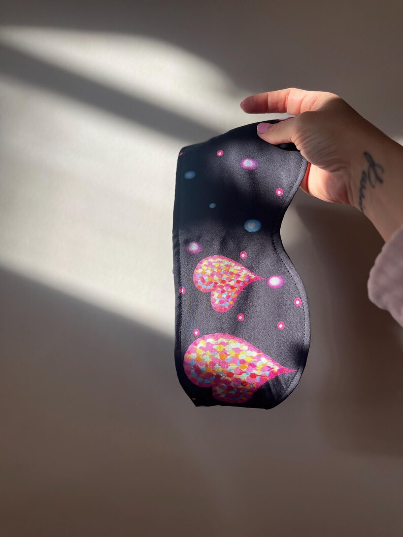 May include: A dark blue sleep mask featuring two heart shapes with colorful, speckled patterns. The mask has a dark background with small, light-colored dots. The mask is held up by a hand.