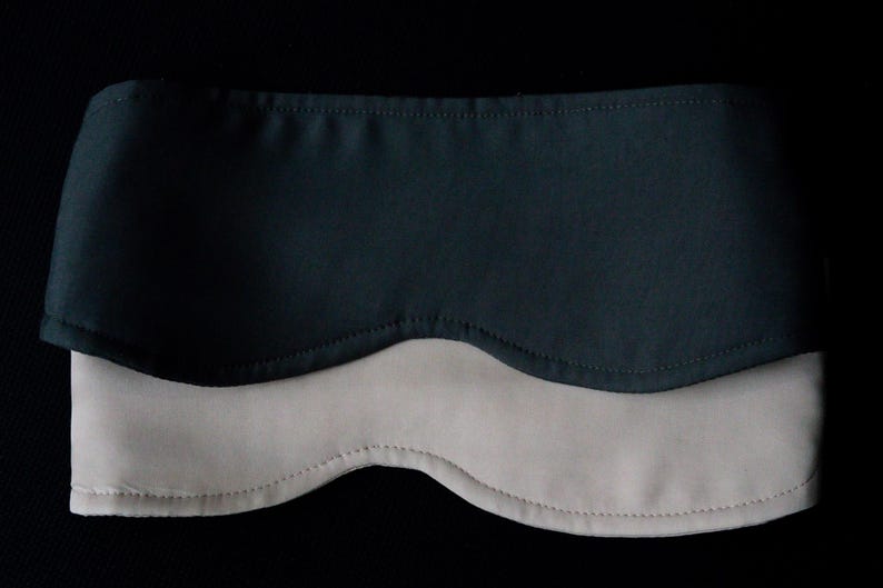 Pu&ograve; includere: Una mascherina per dormire bicolore con una sezione superiore grigio antracite scuro e una sezione inferiore beige chiaro. La mascherina ha una forma curva e sagomata con cuciture visibili lungo i bordi, progettata per coprire gli occhi.