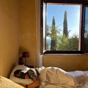K&ouml;nnte beinhalten: Eine Person liegt im Bett, tr&auml;gt eine schwarze Schlafmaske und ist mit einer wei&szlig;en Bettdecke zugedeckt. Der Raum hat gelbe W&auml;nde, eine kleine Lampe mit dunklem Schirm und ein offenes Fenster mit Blick auf Zypressen vor blauem Himmel.
