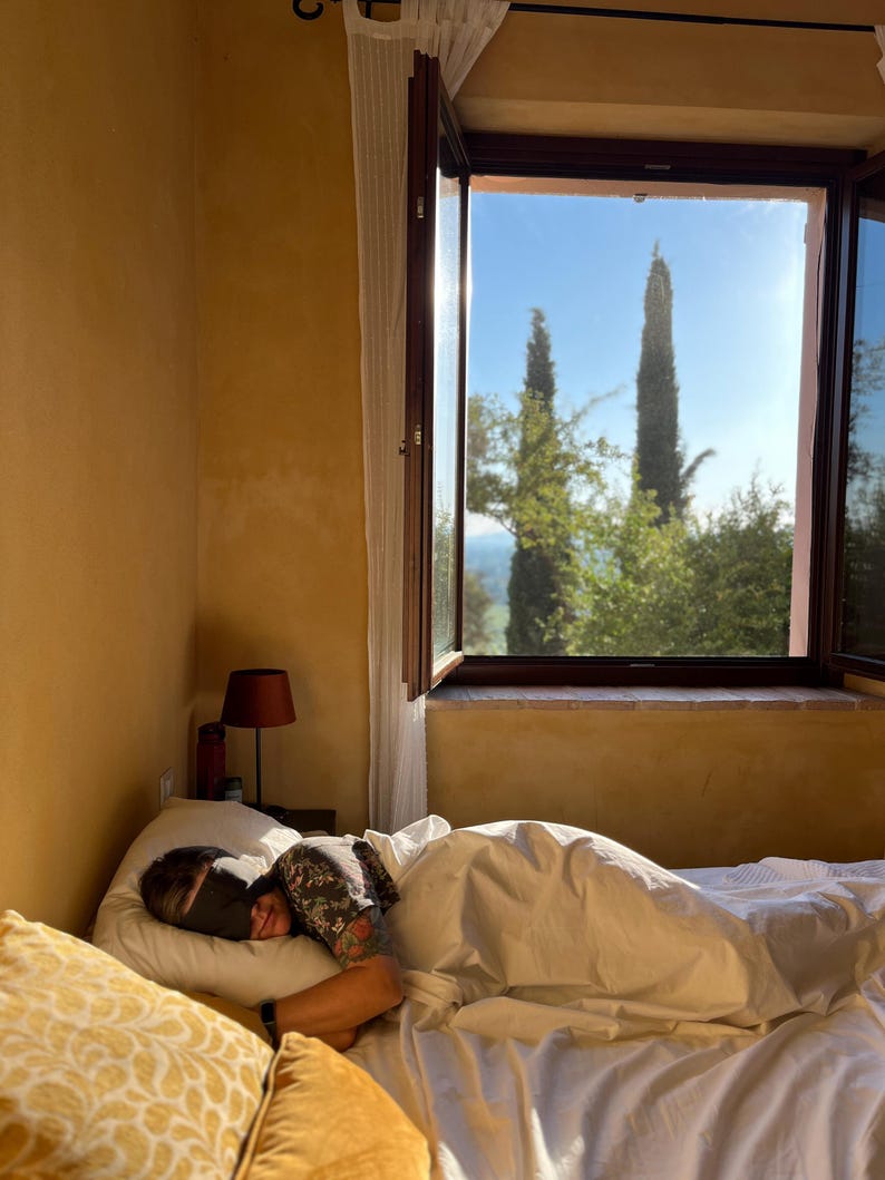 K&ouml;nnte beinhalten: Eine Person liegt im Bett, tr&auml;gt eine schwarze Schlafmaske und ist mit einer wei&szlig;en Bettdecke zugedeckt. Der Raum hat gelbe W&auml;nde, eine kleine Lampe mit dunklem Schirm und ein offenes Fenster mit Blick auf Zypressen vor blauem Himmel.