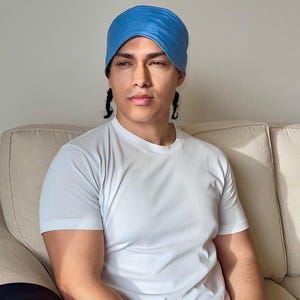 Op de afbeelding: Een lichtblauwe hoofdband wordt gedragen. De persoon draagt een wit t-shirt. De persoon zit op een beige bank.
