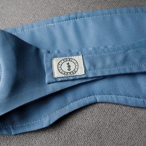 Op de afbeelding: Een lichtblauw stoffen item met witte stiksels en een klein rechthoekig label. Het label heeft de tekst "COSY LONDON" in een cirkelvormig ontwerp. Het item is opgevouwen en lijkt gemaakt van een zacht materiaal.