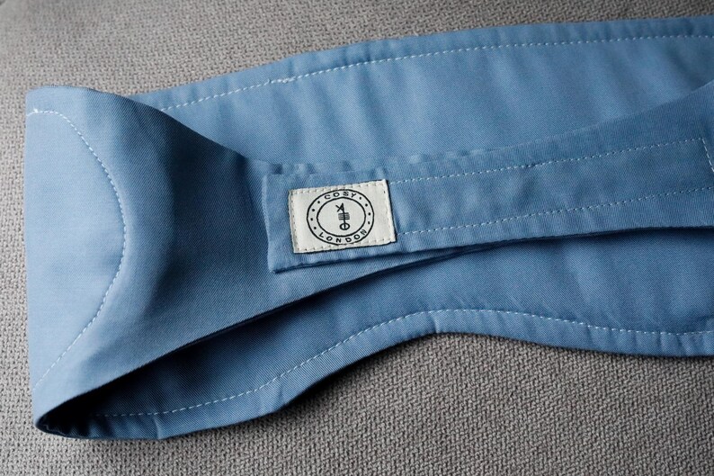 Op de afbeelding: Een lichtblauw stoffen item met witte stiksels en een klein rechthoekig label. Het label heeft de tekst "COSY LONDON" in een cirkelvormig ontwerp. Het item is opgevouwen en lijkt gemaakt van een zacht materiaal.
