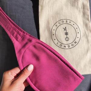 May include: A magenta-colored sleep mask with a stitched edge is held near a beige drawstring bag. The bag has a black circular logo with the words "COSY LONDON" and a symbol. The mask is made of a soft material.
