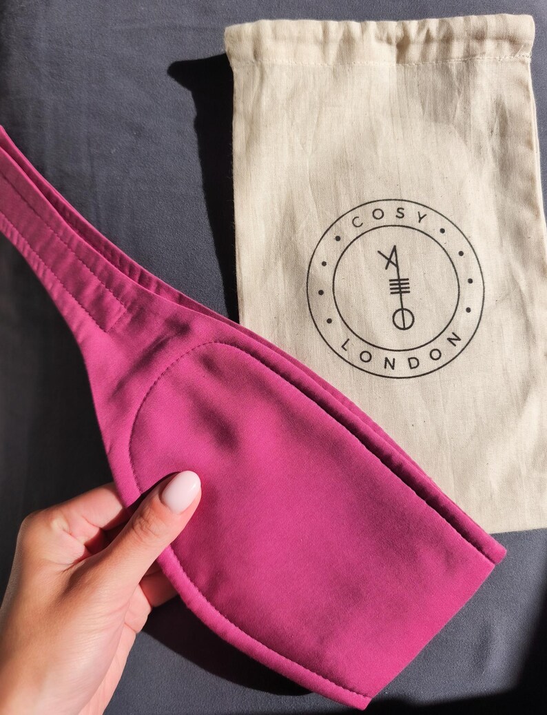 May include: A magenta-colored sleep mask with a stitched edge is held near a beige drawstring bag. The bag has a black circular logo with the words "COSY LONDON" and a symbol. The mask is made of a soft material.
