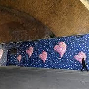 May include: A street art mural under an arched bridge features large pink heart shapes against a dark blue background with white dots. A person walks past the mural on the sidewalk.