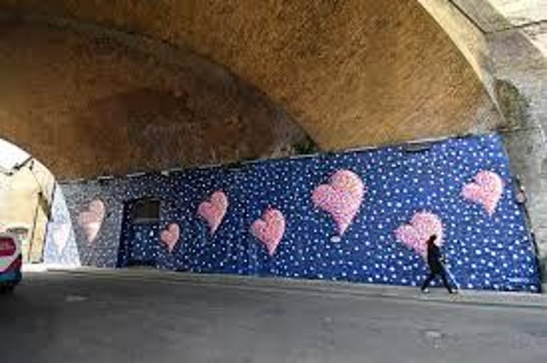 May include: A street art mural under an arched bridge features large pink heart shapes against a dark blue background with white dots. A person walks past the mural on the sidewalk.