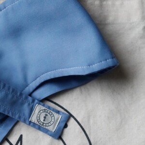 Op de afbeelding: Een lichtblauw stoffen item met witte stiksels, mogelijk een hoed of accessoire, ligt op een canvas draagtas. De tas heeft een zwart grafisch ontwerp en een klein label met tekst. De achtergrond is zwart.