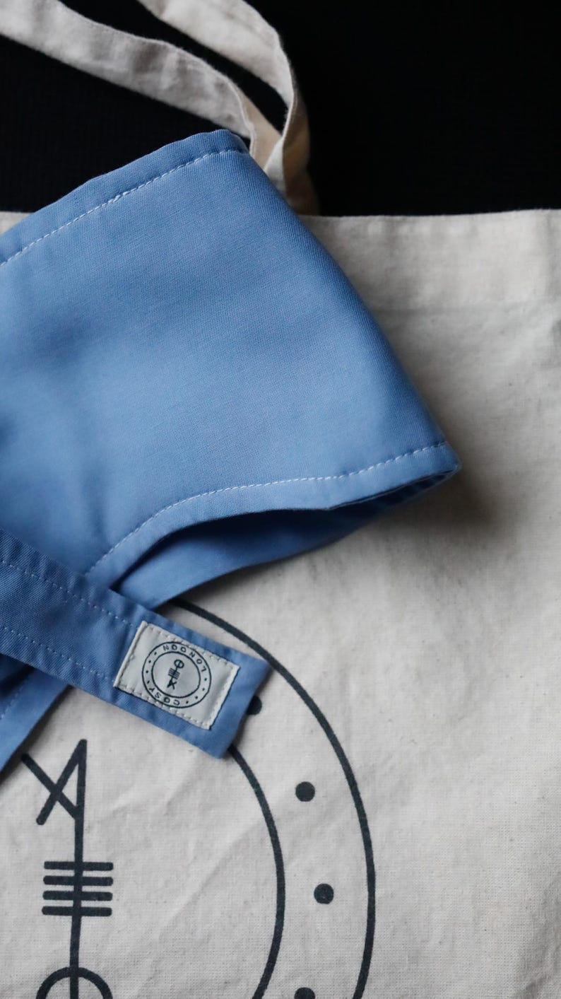 Op de afbeelding: Een lichtblauw stoffen item met witte stiksels, mogelijk een hoed of accessoire, ligt op een canvas draagtas. De tas heeft een zwart grafisch ontwerp en een klein label met tekst. De achtergrond is zwart.