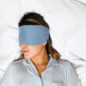 Op de afbeelding: Een persoon met een lichtblauw slaapmasker, liggend op een wit bed. De persoon draagt een blauw-wit gestreept pyjamajasje. Het slaapmasker bedekt de ogen.