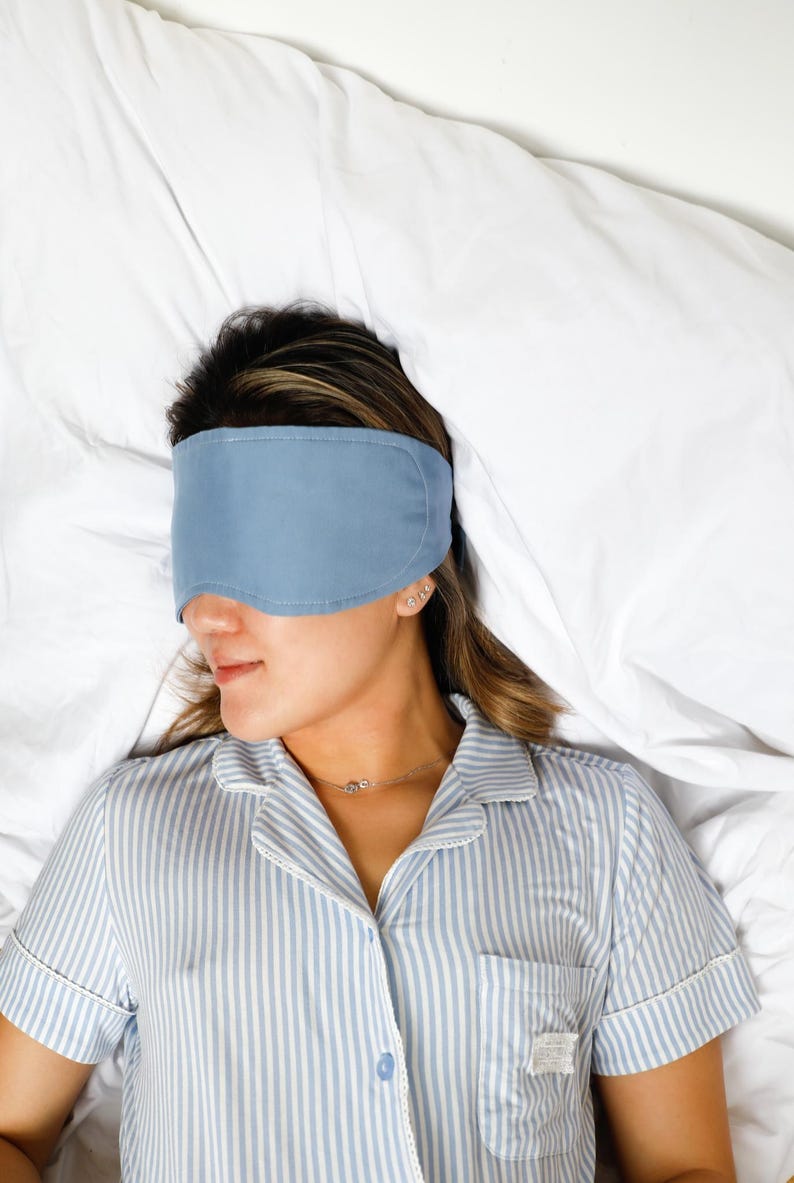 Op de afbeelding: Een persoon met een lichtblauw slaapmasker, liggend op een wit bed. De persoon draagt een blauw-wit gestreept pyjamajasje. Het slaapmasker bedekt de ogen.