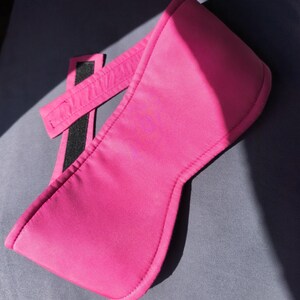May include: A bright pink sleep mask with a wide, curved front and a strap with a black Velcro closure. The mask is made of a smooth, possibly satin-like material, and is designed to block out light.
