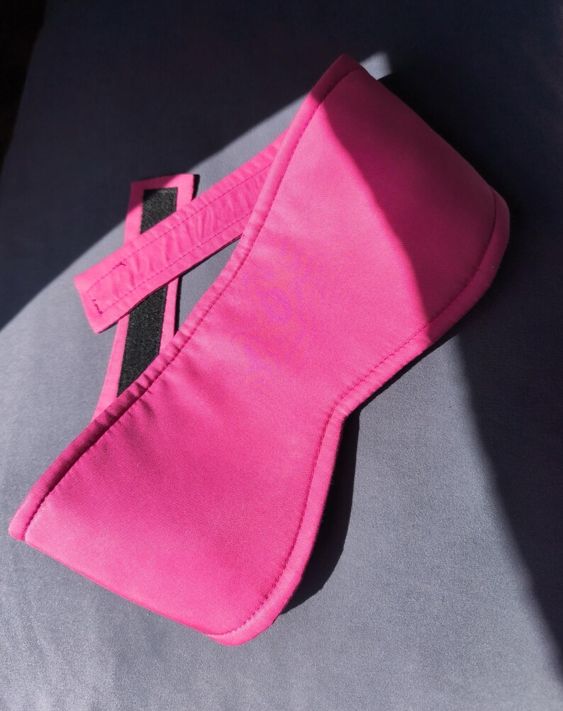 May include: A bright pink sleep mask with a wide, curved front and a strap with a black Velcro closure. The mask is made of a smooth, possibly satin-like material, and is designed to block out light.