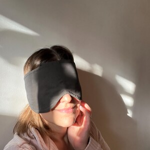 K&ouml;nnte beinhalten: Eine einfarbige, dunkelgraue Schlafmaske, die die Augen bedeckt und eine konturierte Form aufweist, die &uuml;ber den Nasenr&uuml;cken passt und bis zu den Schl&auml;fen reicht. Der Stoff wirkt glatt und blickdicht und dient dazu, Licht f&uuml;r Ruhe zu blockieren.