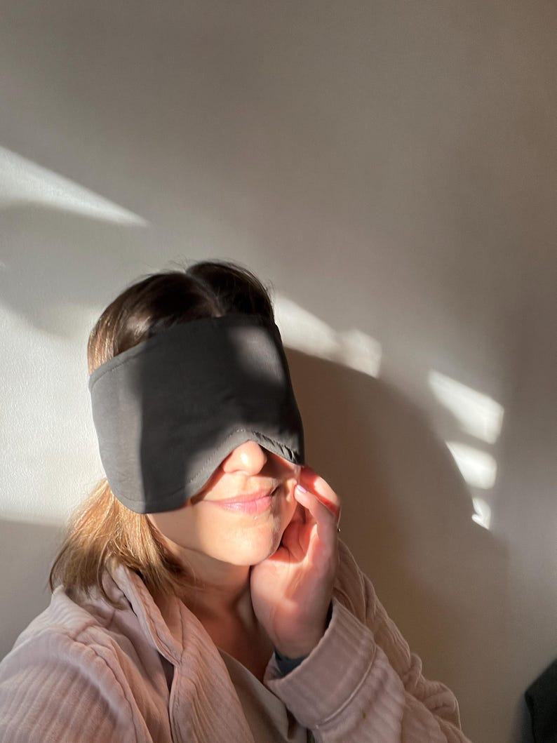 K&ouml;nnte beinhalten: Eine einfarbige, dunkelgraue Schlafmaske, die die Augen bedeckt und eine konturierte Form aufweist, die &uuml;ber den Nasenr&uuml;cken passt und bis zu den Schl&auml;fen reicht. Der Stoff wirkt glatt und blickdicht und dient dazu, Licht f&uuml;r Ruhe zu blockieren.