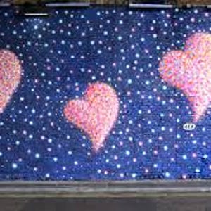 May include: A street art mural features large pink hearts against a dark blue background speckled with white dots. A person in a light-colored coat walks along the sidewalk in front of the mural.