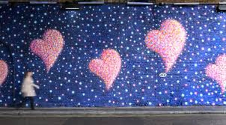 May include: A street art mural features large pink hearts against a dark blue background speckled with white dots. A person in a light-colored coat walks along the sidewalk in front of the mural.