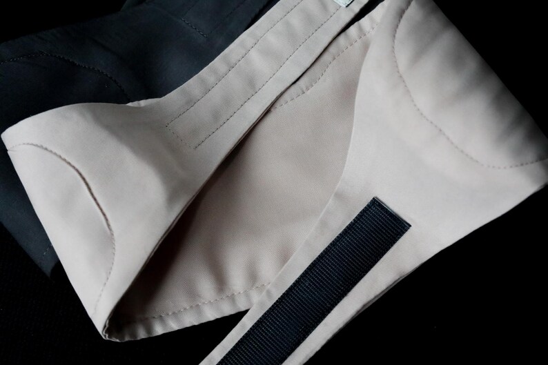 Puede incluir: Primer plano de un cuello de tela beige con una correa negra y detalles de costura. El cuello est&aacute; superpuesto a una tela oscura, creando un contraste. La imagen est&aacute; bien iluminada, destacando la textura y el dise&ntilde;o.