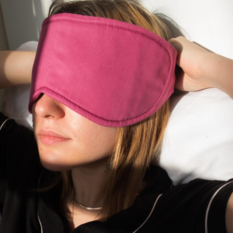 May include: A magenta sleep mask covers the eyes of a person. The mask is made of a soft fabric and has a curved shape to fit the face. The person is wearing a black shirt and a silver necklace. The background is a white pillow.