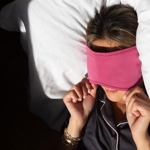 May include: A person wearing a magenta sleep mask, resting on a white bed with a white comforter. The person is wearing a black pajama top with white trim and gold bracelets.