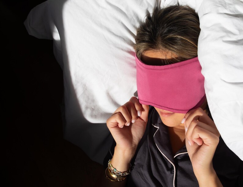 May include: A person wearing a magenta sleep mask, resting on a white bed with a white comforter. The person is wearing a black pajama top with white trim and gold bracelets.