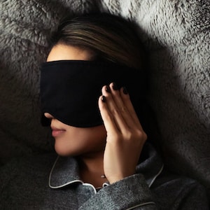 K&ouml;nnte beinhalten: Eine schwarze Schlafmaske bedeckt die Augen einer Person, die auf einer grauen, flauschigen Oberfl&auml;che liegt. Die Person tr&auml;gt ein graues Pyjama-Oberteil mit wei&szlig;er Paspelierung und hat dunklen Nagellack. Die Maske besteht aus weichem, undurchsichtigem Material.