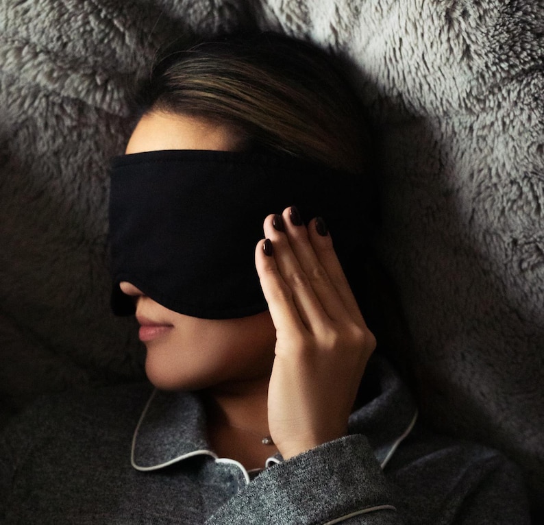 K&ouml;nnte beinhalten: Eine schwarze Schlafmaske bedeckt die Augen einer Person, die auf einer grauen, flauschigen Oberfl&auml;che liegt. Die Person tr&auml;gt ein graues Pyjama-Oberteil mit wei&szlig;er Paspelierung und hat dunklen Nagellack. Die Maske besteht aus weichem, undurchsichtigem Material.