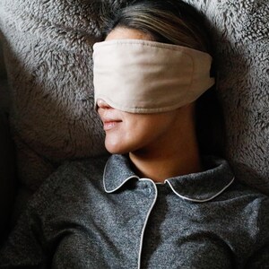 Puede incluir: Una persona con una m&aacute;scara para dormir beige descansa sobre una superficie gris y esponjosa. Lleva una camisa de pijama gris con ribete blanco. La m&aacute;scara para dormir est&aacute; hecha de un material suave, dise&ntilde;ada para la comodidad y la relajaci&oacute;n.