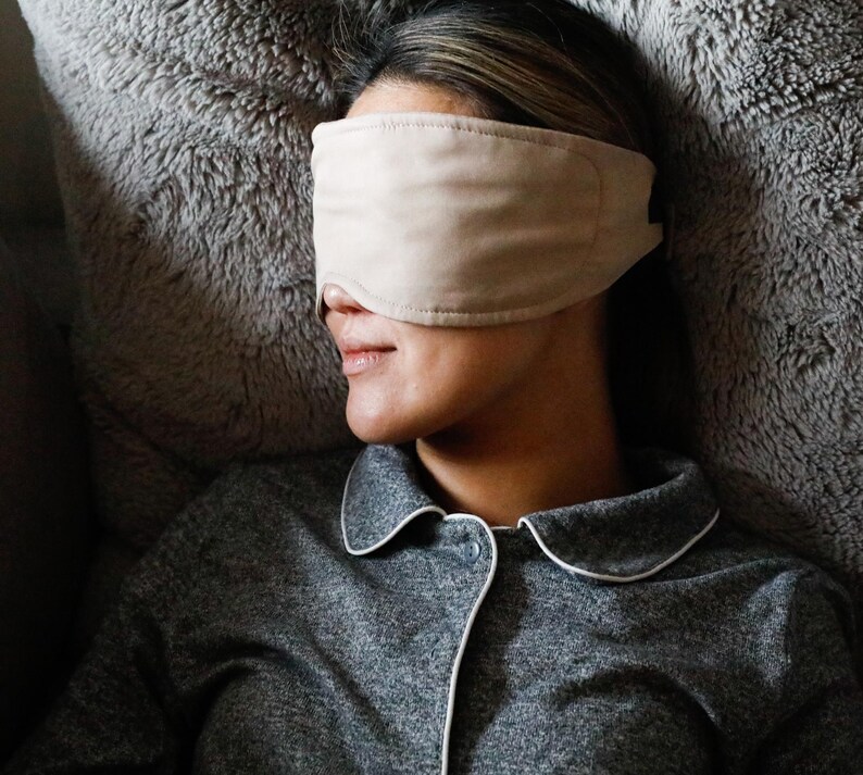 Puede incluir: Una persona con una m&aacute;scara para dormir beige descansa sobre una superficie gris y esponjosa. Lleva una camisa de pijama gris con ribete blanco. La m&aacute;scara para dormir est&aacute; hecha de un material suave, dise&ntilde;ada para la comodidad y la relajaci&oacute;n.