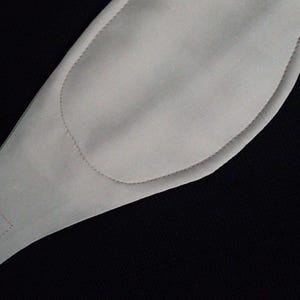 Puede incluir: Una m&aacute;scara para los ojos acolchada de color gris claro, con forma curva y una leng&uuml;eta rectangular. La m&aacute;scara tiene un contorno cosido y se encuentra sobre un fondo negro. La m&aacute;scara parece estar hecha de un material suave, posiblemente tela.
