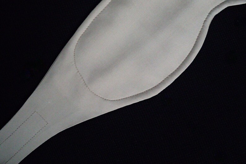 Puede incluir: Una m&aacute;scara para los ojos acolchada de color gris claro, con forma curva y una leng&uuml;eta rectangular. La m&aacute;scara tiene un contorno cosido y se encuentra sobre un fondo negro. La m&aacute;scara parece estar hecha de un material suave, posiblemente tela.