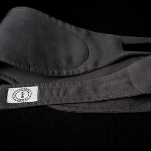 K&ouml;nnte beinhalten: Eine dunkelgraue Schlafmaske mit weichem, gepolstertem Design. Die Maske hat ein rechteckiges Etikett mit Text und einem kreisf&ouml;rmigen Logo. Die Maske besteht aus einem glatten Stoff und ist so konzipiert, dass sie Licht blockiert.