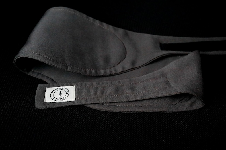 K&ouml;nnte beinhalten: Eine dunkelgraue Schlafmaske mit weichem, gepolstertem Design. Die Maske hat ein rechteckiges Etikett mit Text und einem kreisf&ouml;rmigen Logo. Die Maske besteht aus einem glatten Stoff und ist so konzipiert, dass sie Licht blockiert.