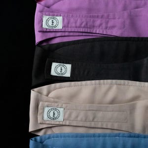 May include: A stack of headbands in various colors: magenta, purple, black, tan, and blue. Each headband has a rectangular label with a circular logo and the text "COSY LONDON". The headbands are made of a smooth fabric.