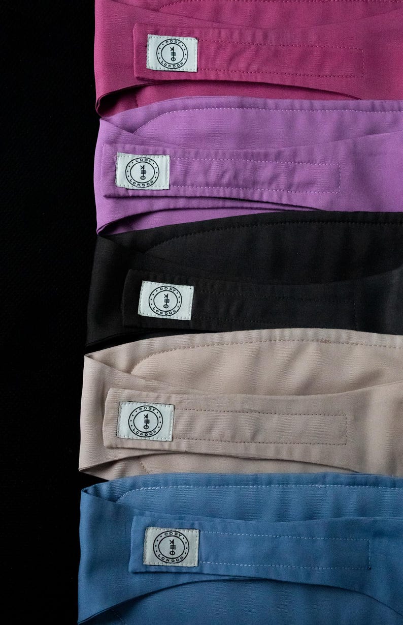 May include: A stack of headbands in various colors: magenta, purple, black, tan, and blue. Each headband has a rectangular label with a circular logo and the text "COSY LONDON". The headbands are made of a smooth fabric.