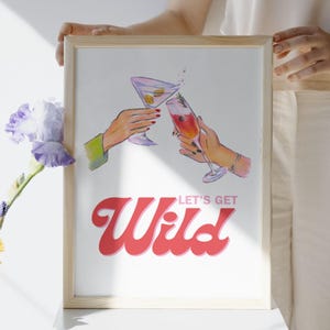 Könnte beinhalten: Ein gerahmter Druck mit einer Aquarellillustration von zwei Händen, die mit Cocktails anstoßen. Eine Hand hält einen Martini mit Oliven, die andere einen rosa Cocktail. Die Worte "LET'S GET Wild" sind in roter Schrift.