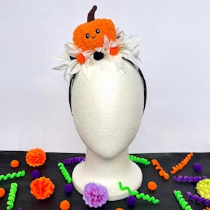 Puede incluir: Una diadema negra con una calabaza de peluche naranja con tallo marrón y cara sonriente, rodeada de pétalos de margarita artificiales blancos y pequeños pompones naranjas y negros. La calabaza tiene ojos y una boca bordados en negro.