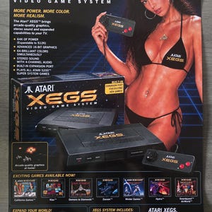 Puede incluir: Un póster publicitario vintage del sistema de videojuegos Atari XEGS que muestra la consola negra, un mando rectangular con dos botones rojos y la caja. El texto destaca las especificaciones técnicas y los títulos de juegos.
